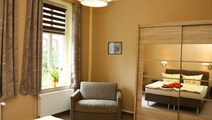 Een slaapkamer met een bed, een stoel, een raam met gordijnen, een radiator en een spiegel.