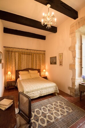 A bedroom with a large bed, wooden beams, a chandelier, and a stone wall.