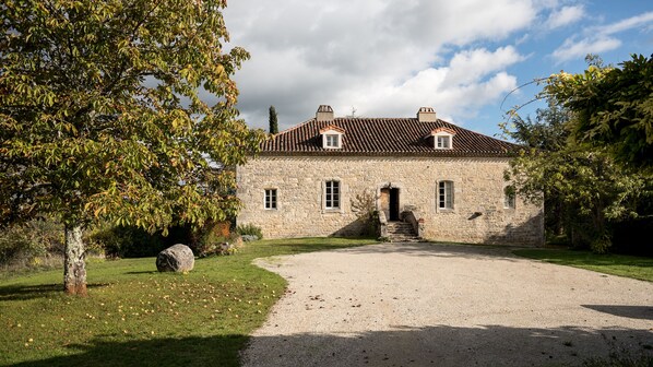 A stone building with a tiled roof, a gravel driveway, and a large tree in the foreground.