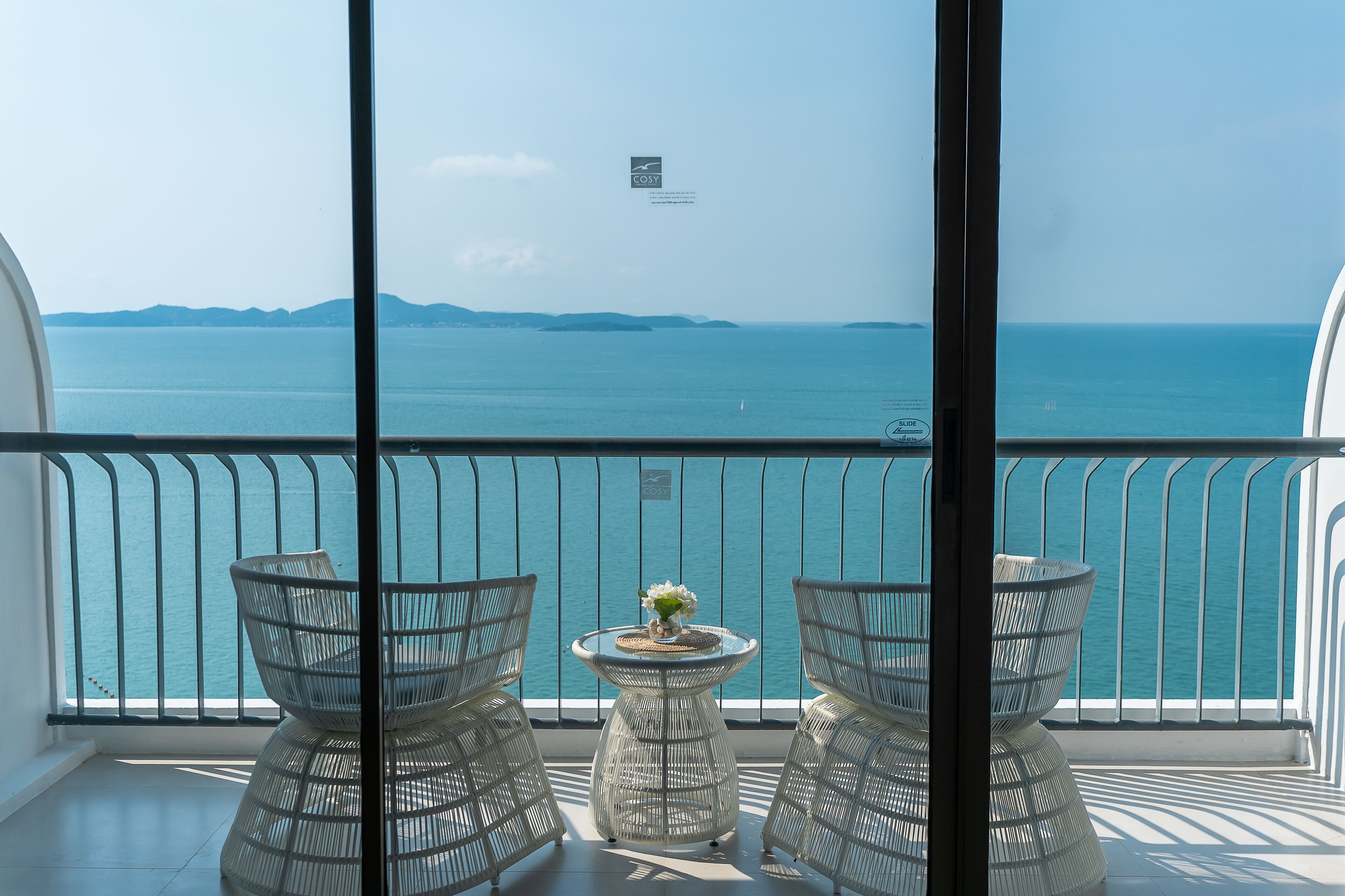 A balcony with two wicker chairs and a small table, overlooking a calm sea with distant landforms.