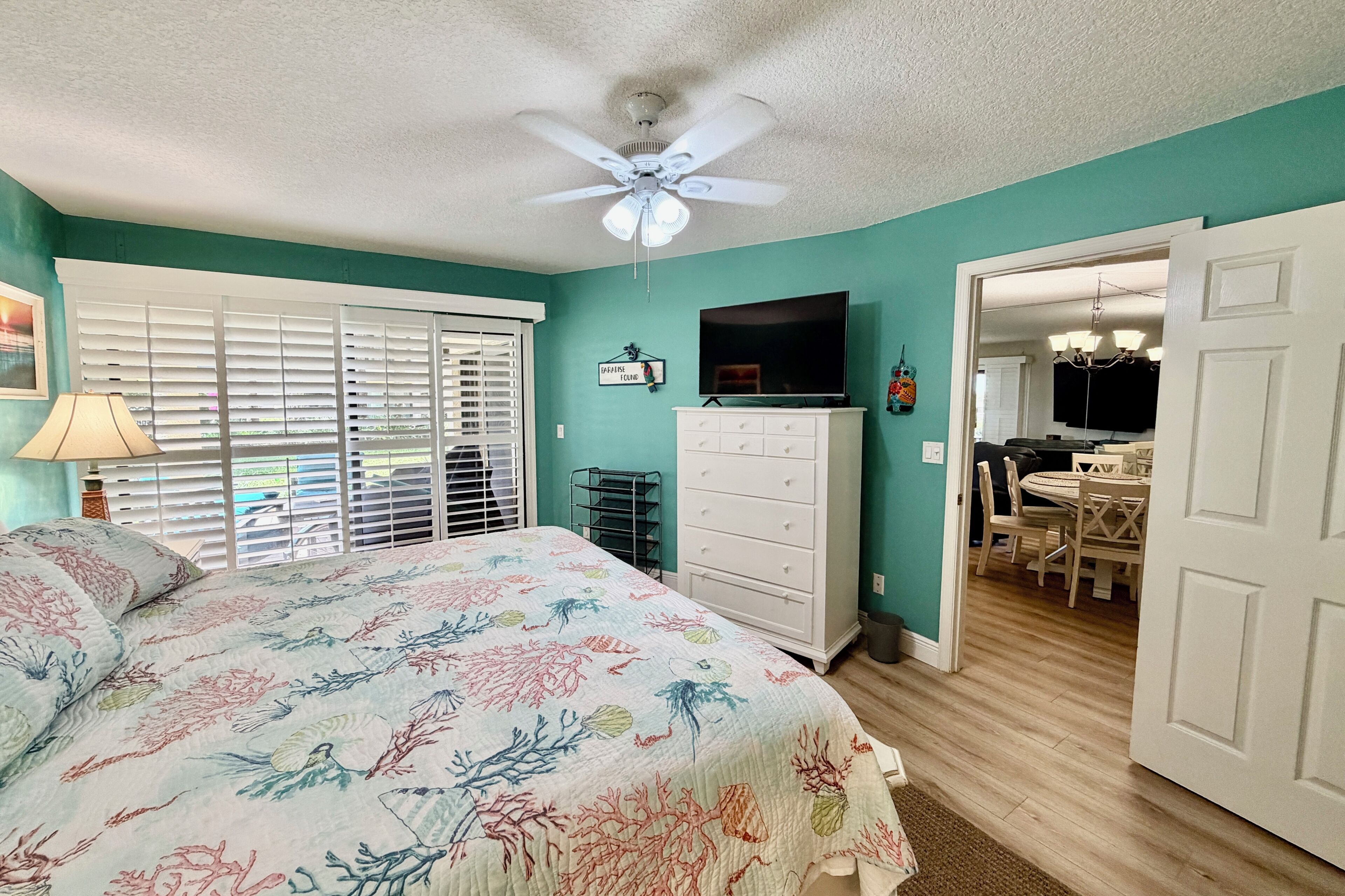 A bedroom with a bed, a ceiling fan, a dresser, a TV, and a dining area with a table and chairs.
