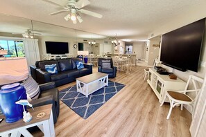 A modern living room with a sofa, coffee table, and TV. There is a dining area with chairs and a table.