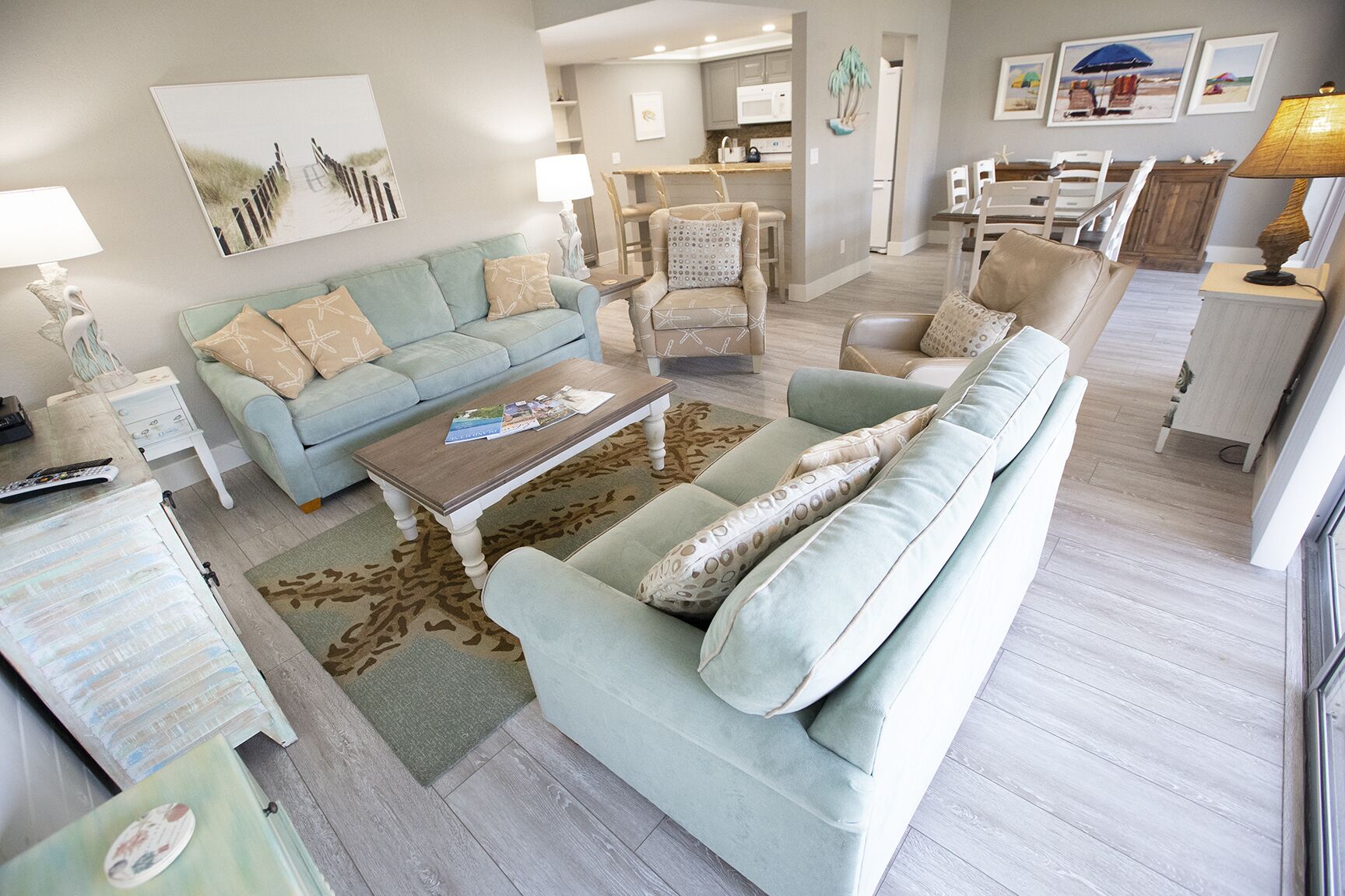 Een ruime woonkamer met een lichtblauwe bank, een bijpassende fauteuil en een salontafel. Op de achtergrond is een keuken te zien met een eettafel en stoelen.
