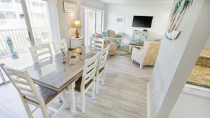 Un coin repas avec une table à plateau en verre et des chaises blanches, un canapé et un téléviseur dans le salon.
