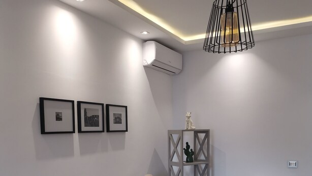 Een moderne woonkamer met een bank, een salontafel en een stoel. Er hangen ingelijste foto's aan de muur, een decoratief object op een plank en staat een airconditioner.