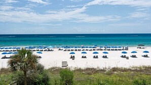 A beach with rows of sun loungers and blue umbrellas, a clear sky, and the ocean in the background.