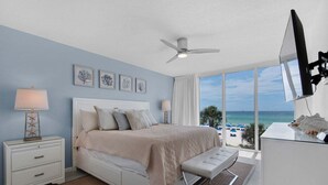 A bedroom with a bed, bedside tables, a ceiling fan, a TV, and a view of the beach.