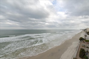 A beach with waves, a cloudy sky, and a building with a fence and palm trees.