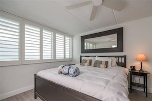 A bedroom with a bed, a nightstand, a lamp, a ceiling fan, and a large window with blinds.