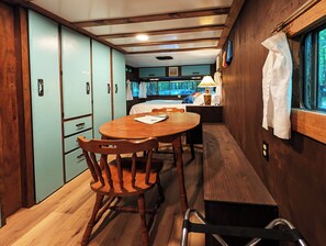 A compact, well-lit room with a wooden dining table, chairs, and a bench. There is a bed with a window view, a small table with a lamp, and a coat rack. - Luxury Glamping w/Resort Access Included, Site 3 (Phelps)