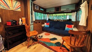 Un intérieur de camping-car accueillant, avec un plancher en bois, un canapé bleu agrémenté de coussins colorés, une table en bois et un petit coin cuisine équipé d’une radio et de produits de nettoyage.