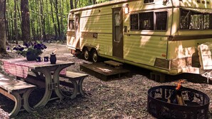 Une vieille caravane est garée dans une forêt, à proximité de tables de pique-nique et d’un foyer de camp.