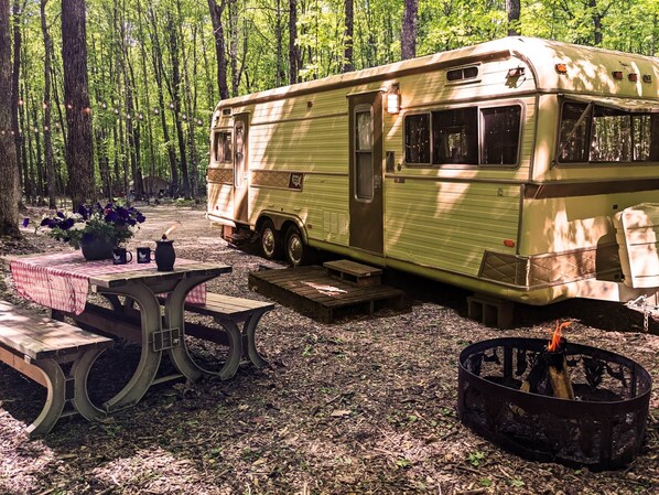 A vintage camper parked in a forest with picnic tables and a campfire. - Luxury Glamping w/Resort Access Included, Site 3 (Phelps)