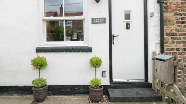 A white building with a black door and a window. There are two potted plants on the ground. - Ascot Cottage (York)