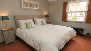A bedroom with a large bed, two bedside tables, a nightstand, a window with wooden shutters, and a radiator. - Ascot Cottage (York)