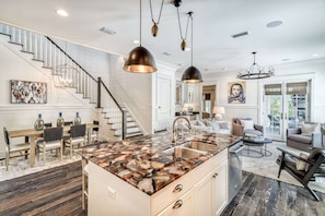 Une cuisine spacieuse dotée d’un îlot central, un coin repas avec une table et des chaises en bois, un escalier et un salon avec un canapé et des fauteuils.