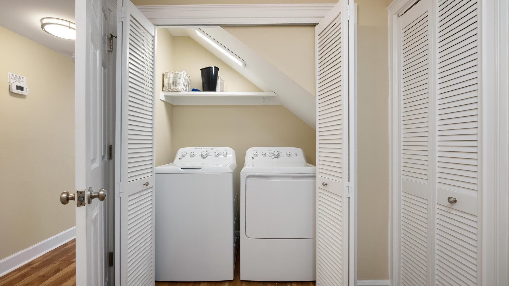 Une buanderie équipée de deux machines à laver et deux sèche-linge à chargement frontal, d’une étagère contenant des produits de lessive et d’un thermostat fixé au mur.