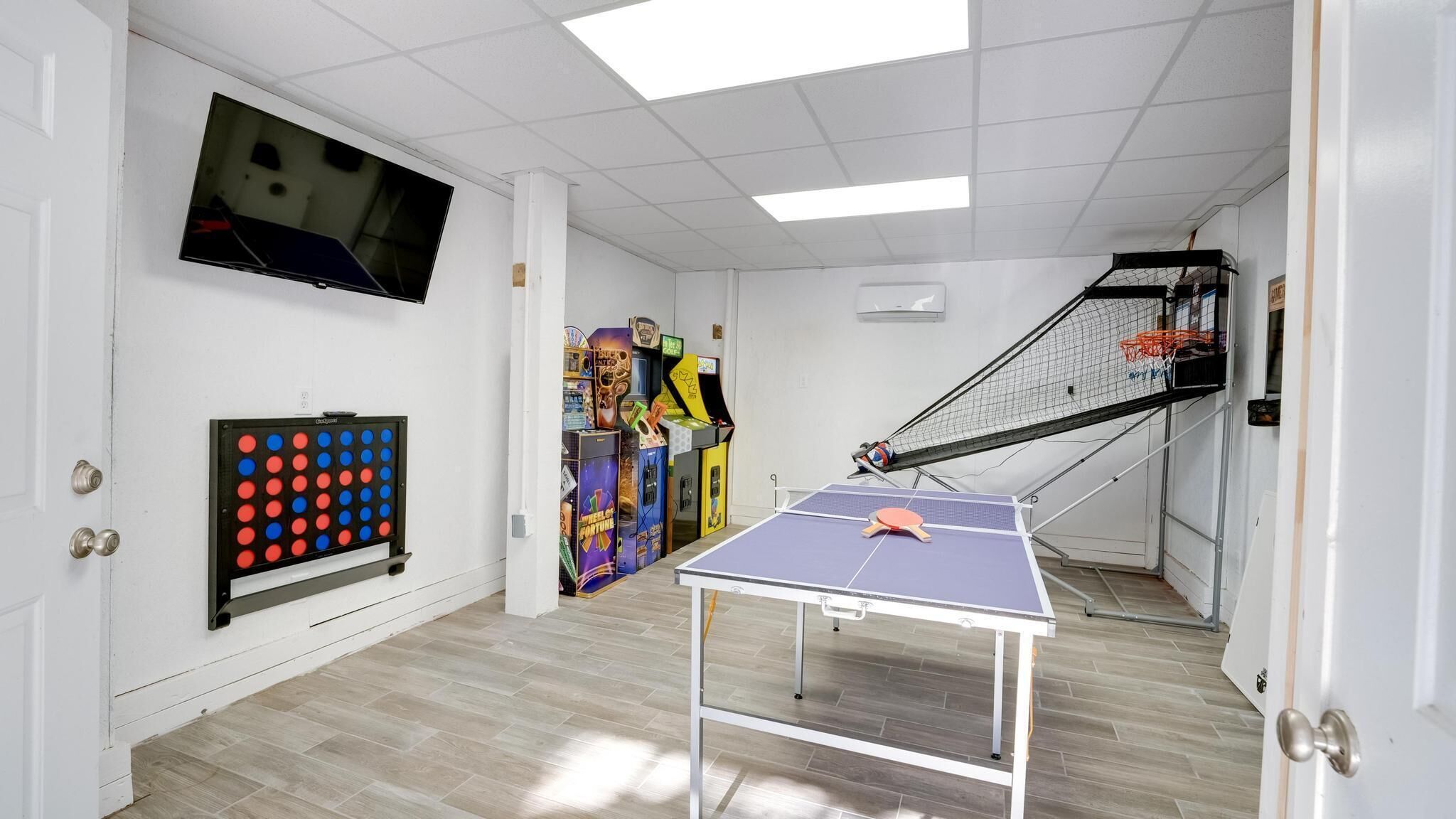 A room with a ping pong table, a TV mounted on the wall, and a variety of arcade games.