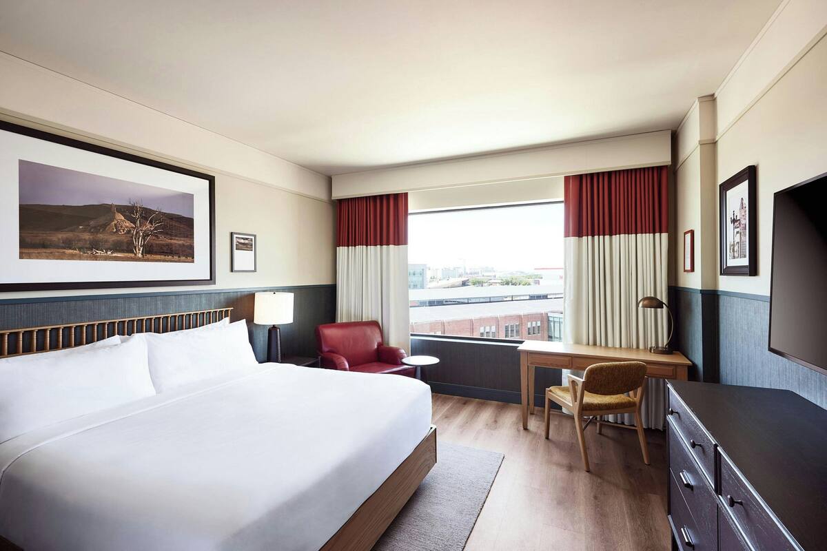 A hotel room with a large bed, a desk, a chair, a TV, and a window with curtains.
