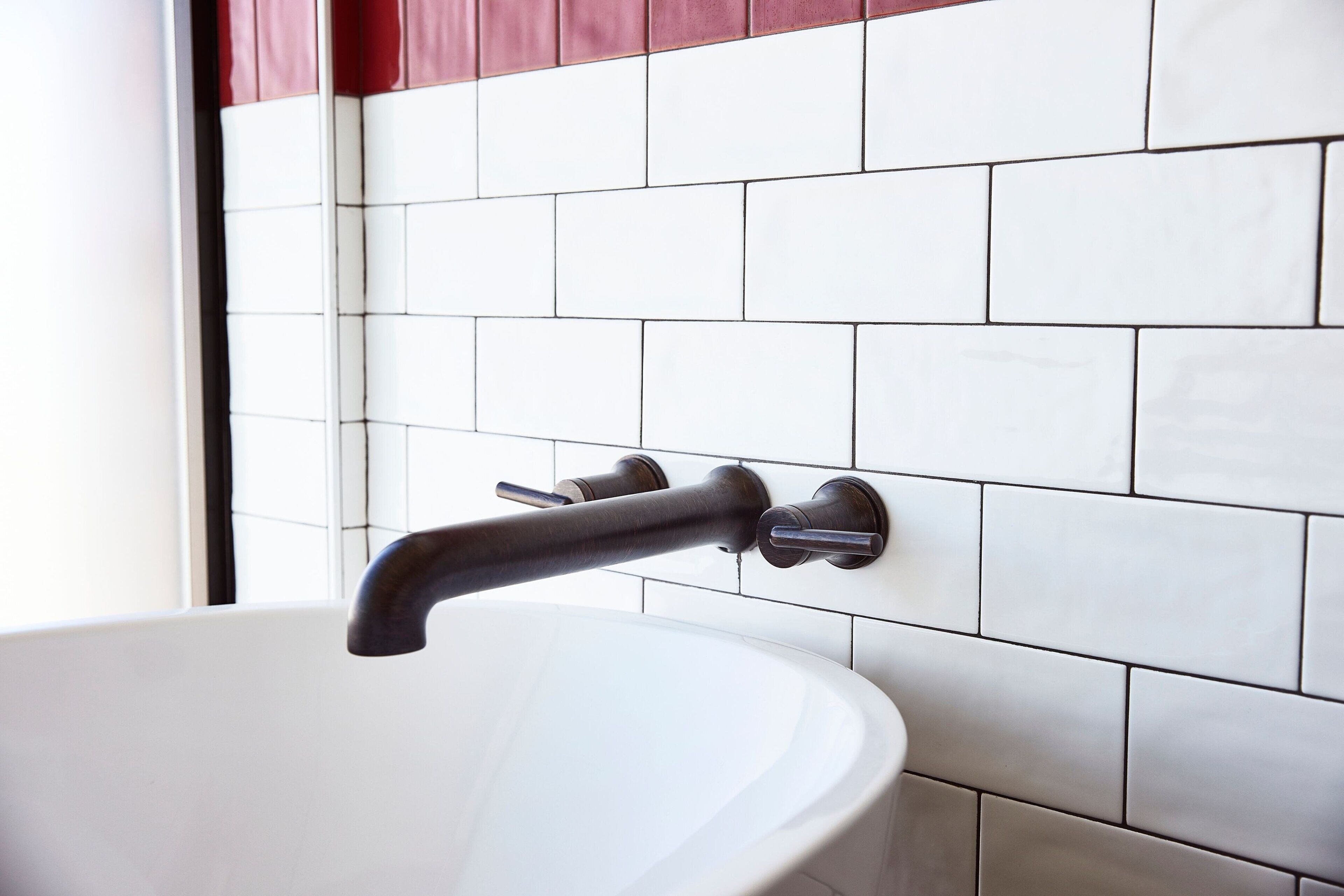 A bathroom with a white sink, a dark faucet, and a tiled wall with a red accent.