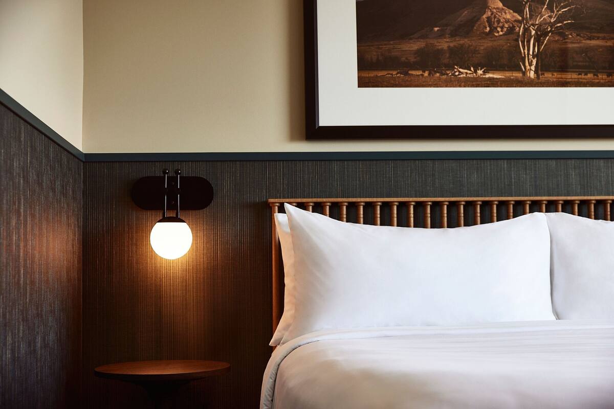 A neatly made bed with white linens, a wooden headboard, a round wooden nightstand, and a wall-mounted light fixture.