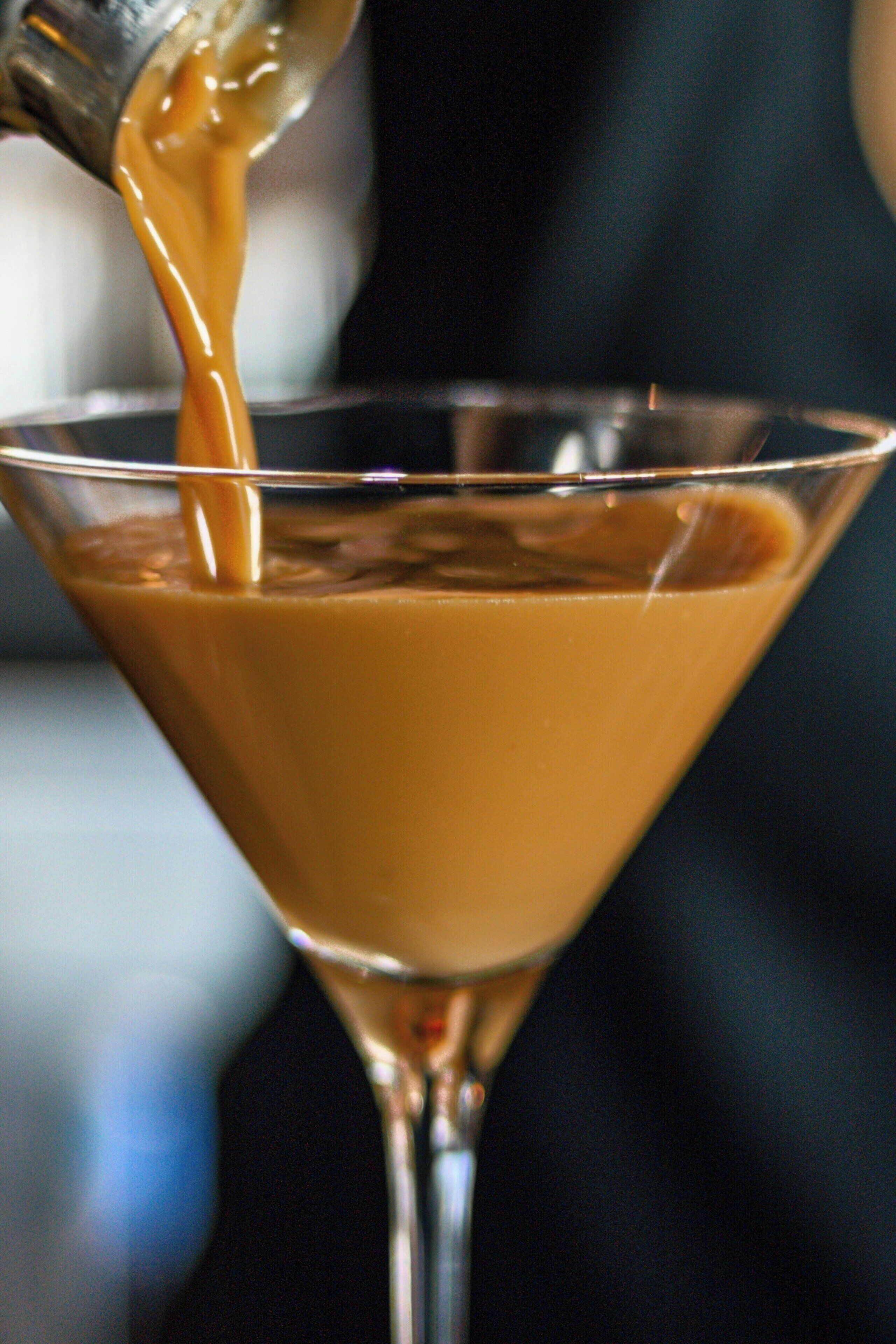 A clear glass martini glass filled with a brown liquid, likely a cocktail, being poured from a metal container.