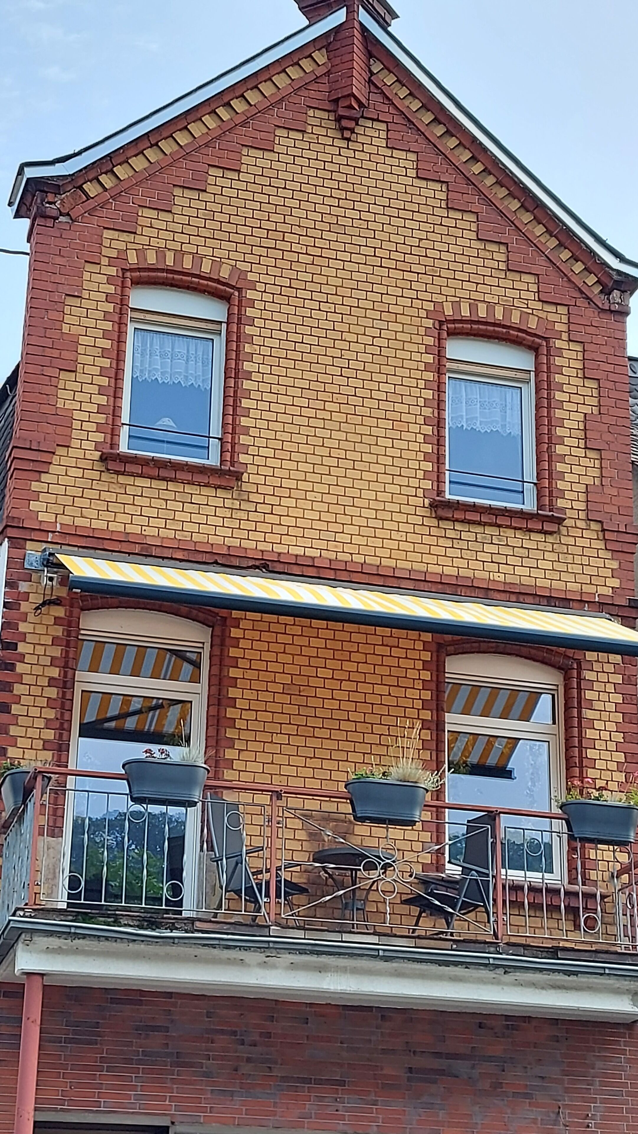 Une maison à deux étages, construite en briques, dotée d’un balcon, d’une terrasse couverte et de deux fenêtres.
