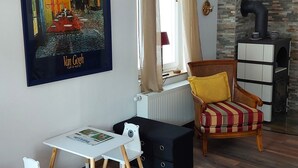 Ein Wohnzimmer mit einem weißen Tisch, einem weißen Sessel, einem hölzernen Stuhl mit gelbem Kissen, einer schwarzen Kommode, einem Kamin und einem Gemälde einer Café-Szene.