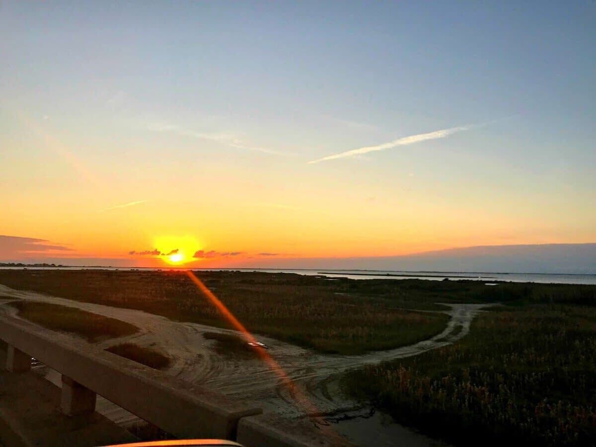 A sunset over a coastal landscape with a dirt road and grassy fields.