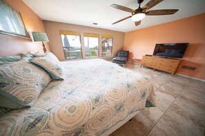 A bedroom with a large bed, a ceiling fan, a TV, and a view of the outdoors.