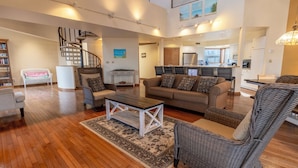 A spacious living area with a wooden floor, a sofa, a coffee table, and a wicker chair. A staircase leads to an upper level. - Big Hilltop Home with Gorgeous Lake Views & Beach Access (Michigan City)