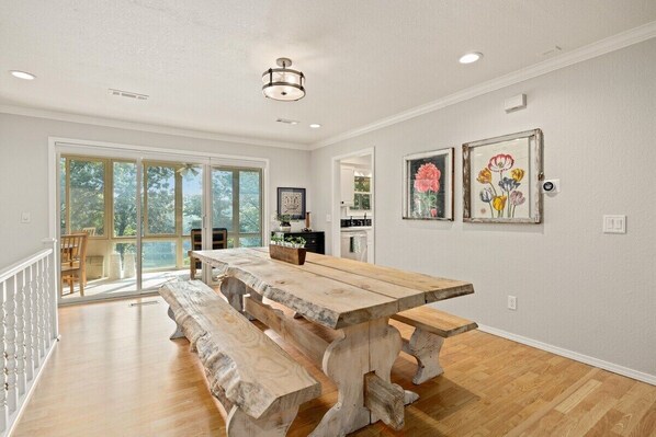 A spacious dining area with a large wooden table and benches, a view through glass doors, and artwork on the walls.