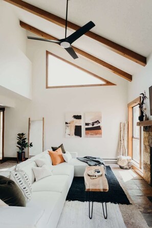 Uma sala de estar espaçosa com teto alto, vigas aparentes, um sofá branco, um ventilador de teto preto e arte abstrata nas paredes.