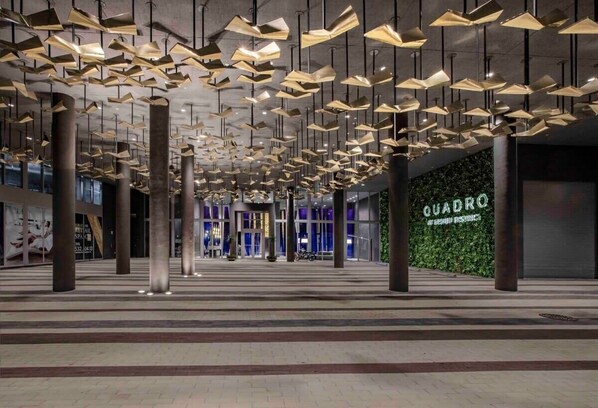 Un vestíbulo amplio con moderna iluminación colgante, pilares grandes y una tienda visible llamada 'QUADRO'.