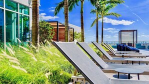Un área de lounge en la azotea con sillones de sol y pequeñas mesas, rodeada de palmeras y un edificio moderno con amplios ventanales.