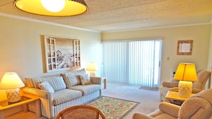 A living room with a striped sofa, a wooden coffee table, and two armchairs. There are lamps on side tables, a mirror on the wall, and a window with blinds. - Barefoot Country 304 1 Bedroom Condo by RedAwning (Ocean City)