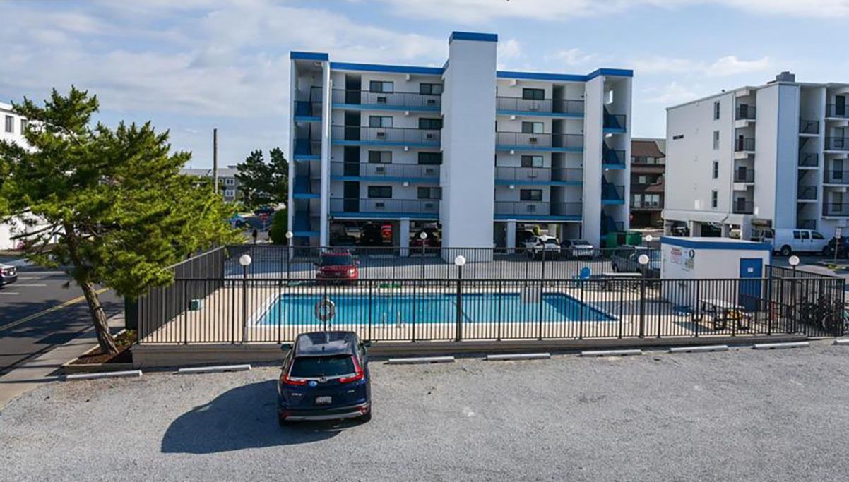 Un edificio residencial de varios pisos con piscina, un estacionamiento cercado y un árbol en primer plano.