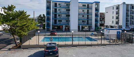 Un edificio residencial de varios pisos con piscina, un estacionamiento cercado y un árbol en primer plano.