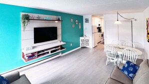 A modern living room with a flat-screen TV, a dining area with white chairs, and a kitchen in the background.