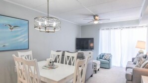 A modern living room with a dining area, a ceiling fan, a flat-screen TV, and a large window with curtains. - Direct ocean front  * 2 Bedrooms * 2 Full Baths + Den * 4th Floor Sleeps 8 * (Ocean City)