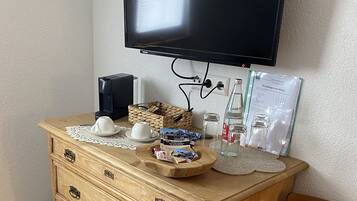 Ein hölzerner Kommode mit Schubladen, ein an der Wand befestigtes Fernsehgerät, ein Sessel mit Kissen und diverse Gegenstände auf der Kommode, darunter eine Kaffeemaschine, eine Flasche und verpackte Waren.