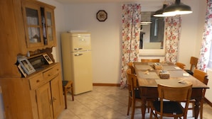 A kitchen with wooden cabinets, a refrigerator, a dining table with chairs, and floral curtains. - s Moose Hof (Weisweil)