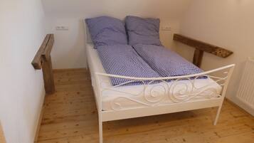 A small, white metal bed with a checkered blue and white bedspread and pillows, positioned in a corner of a room with wooden flooring and white walls.