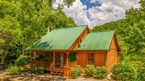 Uma cabana rústica de toras com telhado de metal verde, varanda de madeira e alicerce de pedra, cercada por vegetação exuberante.