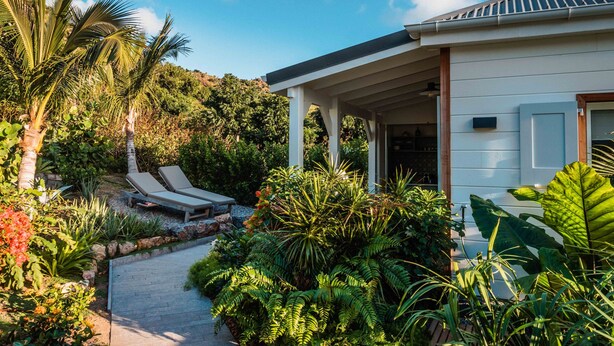 Une maison moderne dotée d’un porche abrité, d’une végétation tropicale luxuriante et d’un chemin menant à un espace de détente avec des chaises longues.