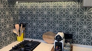 Une cuisine avec un crédence ornée d’un motif géométrique, une cuisinière à gaz, une machine à café et des ustensiles rangés dans un porte-ustensiles jaune.
