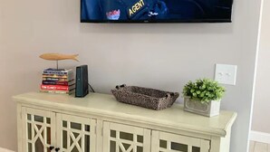 A television mounted on a wall displaying a scene with a person labeled "AGENT". Below the TV, there is a white cabinet with decorative cutouts and a basket on top. - Capital City Condo #3 (Frankfort)