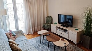 A modern living room with a sofa, a television, a small table, a chair, and a large window with curtains. - Fairytale, Nilie Hospitality MGMT (Thessaloniki)