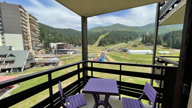 A balcony with two purple chairs and a table, overlooking a playground and a mountainous landscape. - Griou N°52 (Le Lioran)