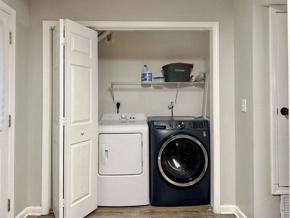 A laundry room with a white washer and dryer, a shelf with laundry supplies, and a door leading to another room.